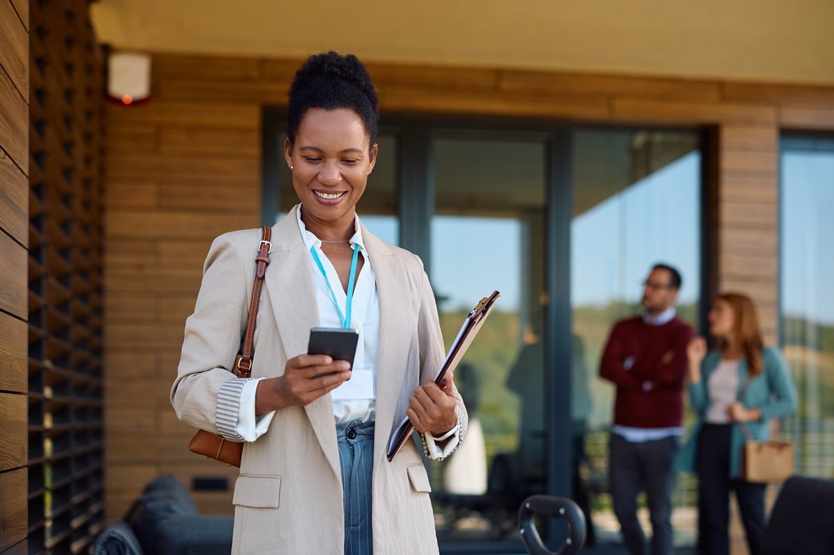 Happy African American real estate agent using cell phone. Her clients are in the background.
