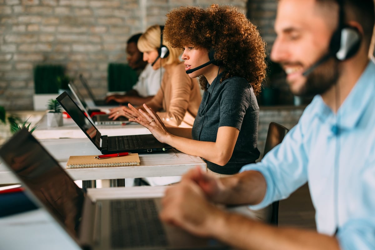 Engaged female customer service agent actively communicates with expressive hand gestures while working on a laptop in a modern, diverse call center