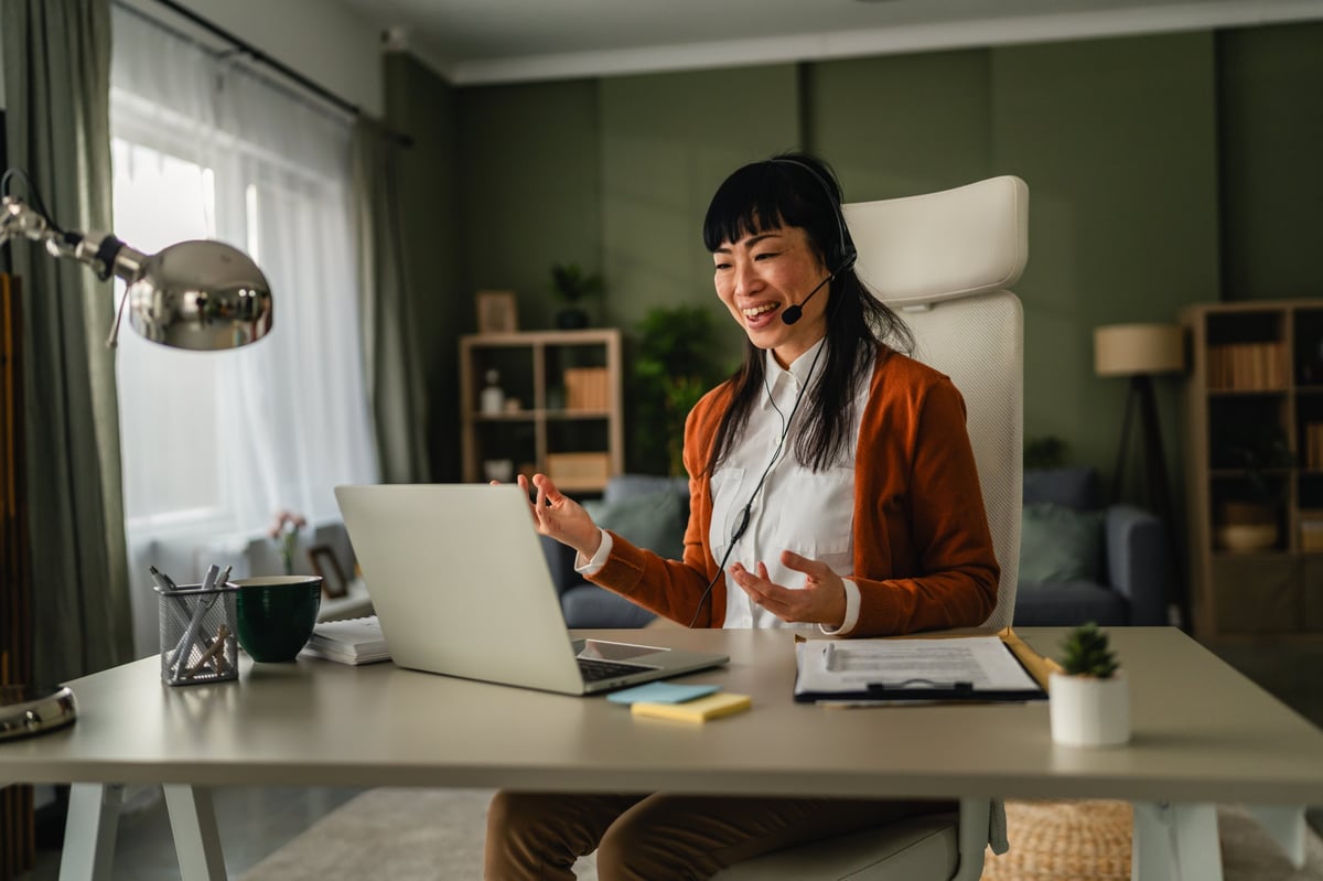 Woman wearing a headset and smiling while typing on her laptop, conducting a virtual meeting or providing customer support from a home office workspace