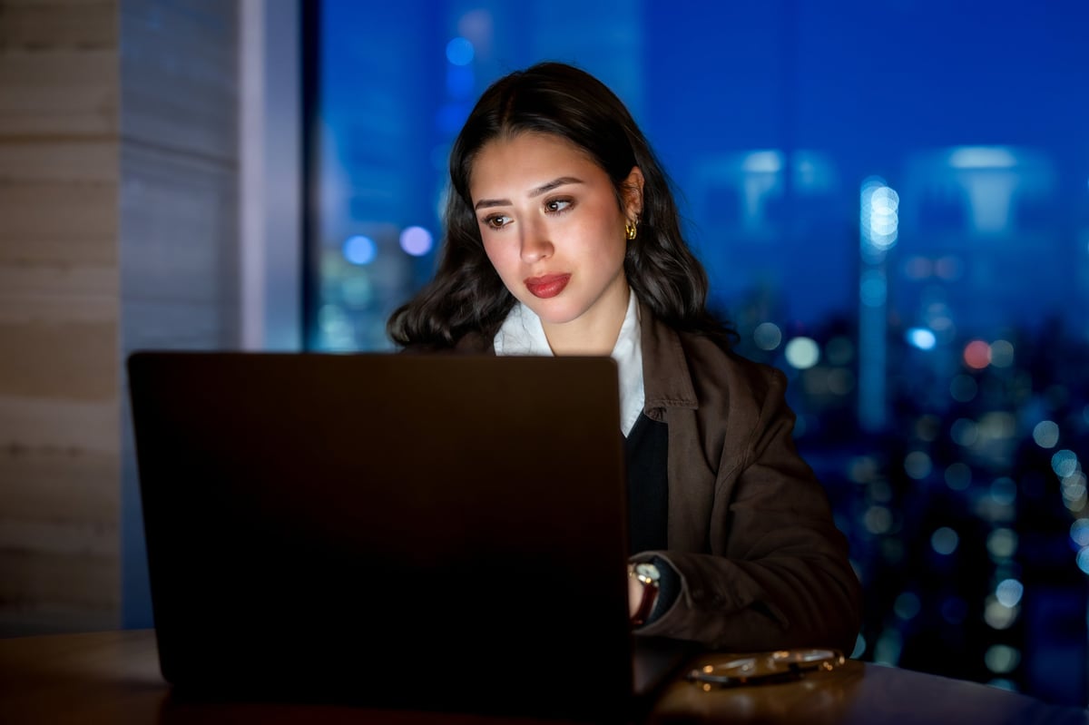 Young adult businesswoman concentrating on her laptop, working late at an office in manhattan with city lights in the background, representing dedication and success