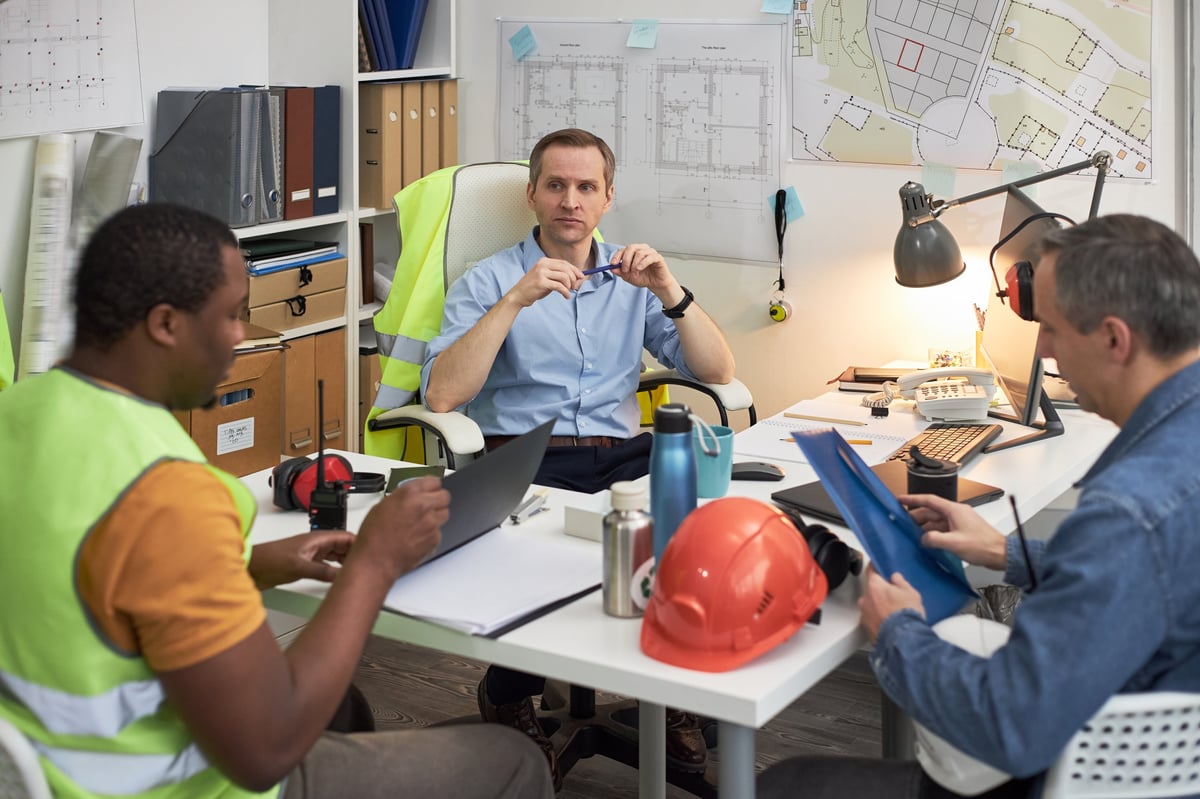 Caucasian middle aged man sitting at desk leading meeting with Black young adult man and Caucasian middle aged man in office, discussing construction plans and reviewing documents