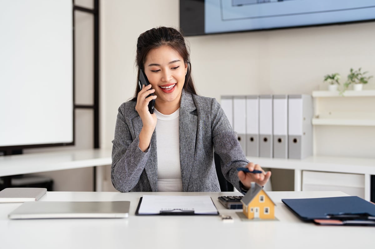 Confident asian business lady, broker real estate agent or manager, sitting at table in a modern office, working with laptop, talking to client or employees on smartphone, smiling.