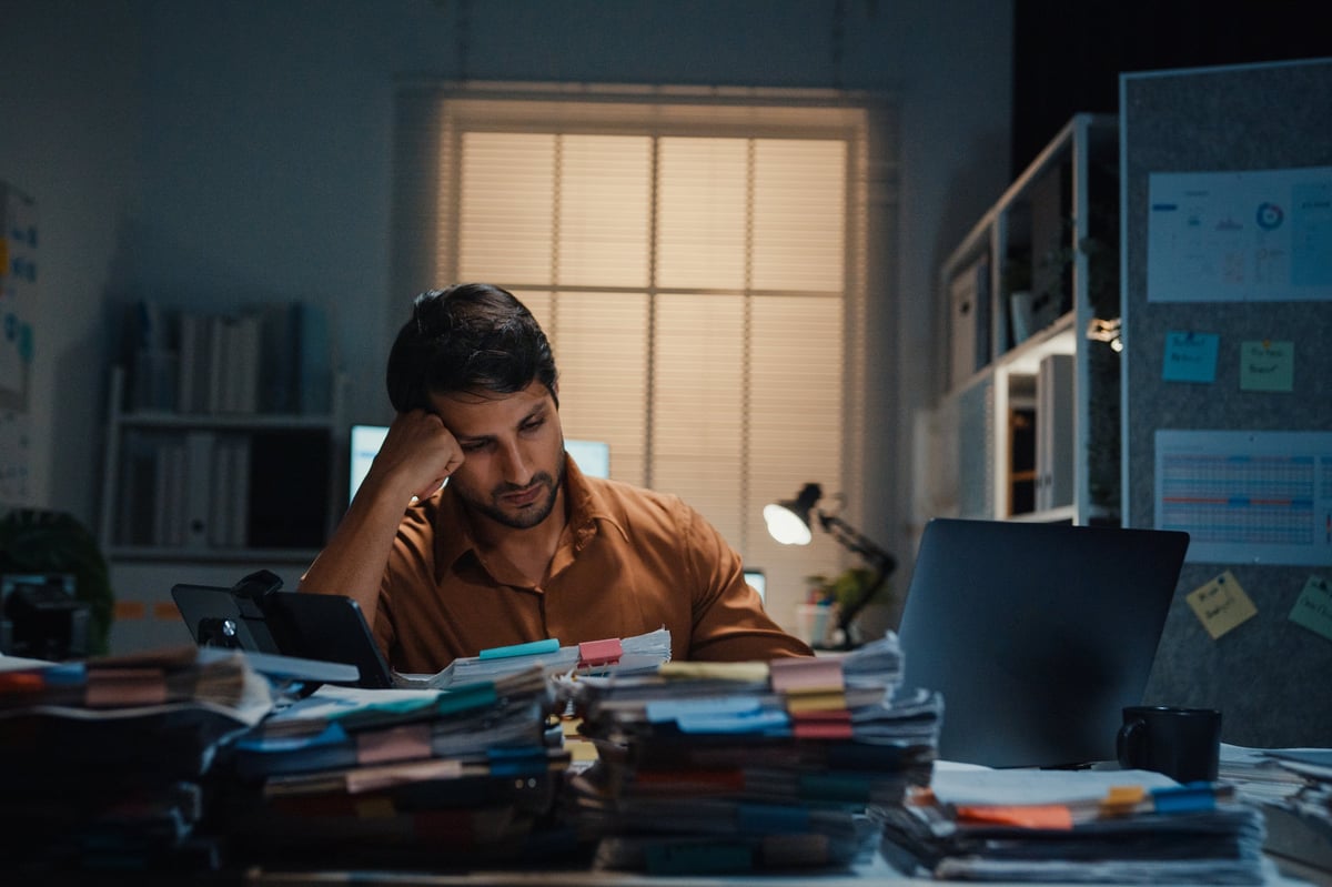 Stressed young Latin businessman having severe headache and migraine at desk in night office.