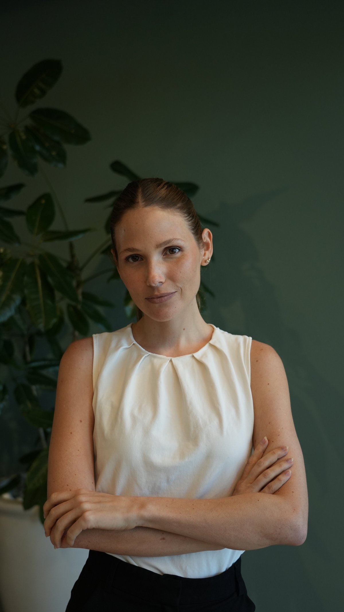 Woman in cream sleeveless top standing with crossed arms against green wall with plant shadow